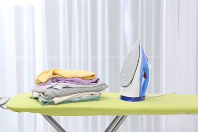 Photo of Board with modern iron and stack of clothes indoors. Laundry day