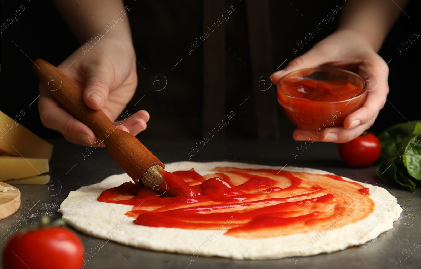 Photo of Woman spreading tomato sauce onto pizza crust at grey table, closeup