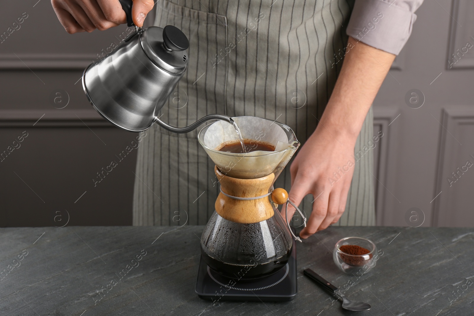 Photo of Man pouring hot water into glass chemex coffeemaker with paper filter and coffee at gray table, closeup