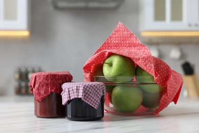 Jars of jams and apples in bowl covered with beeswax food wrap on white table indoors
