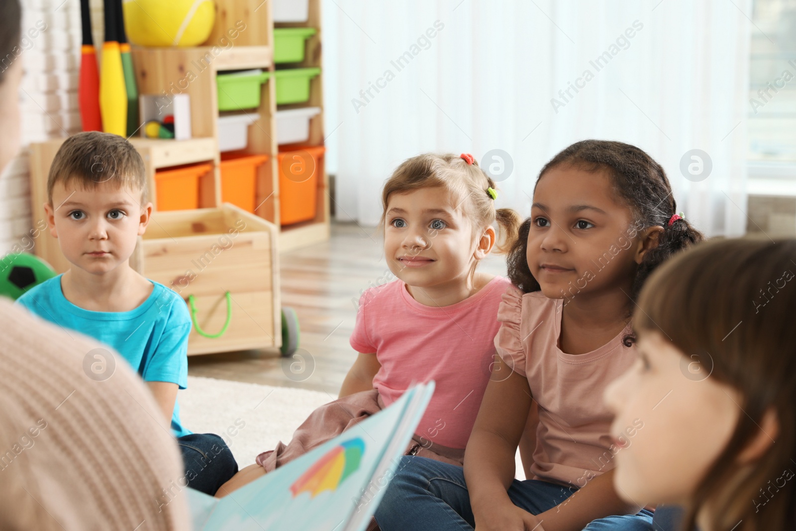 Photo of Kindergarten teacher reading book to cute little children indoors