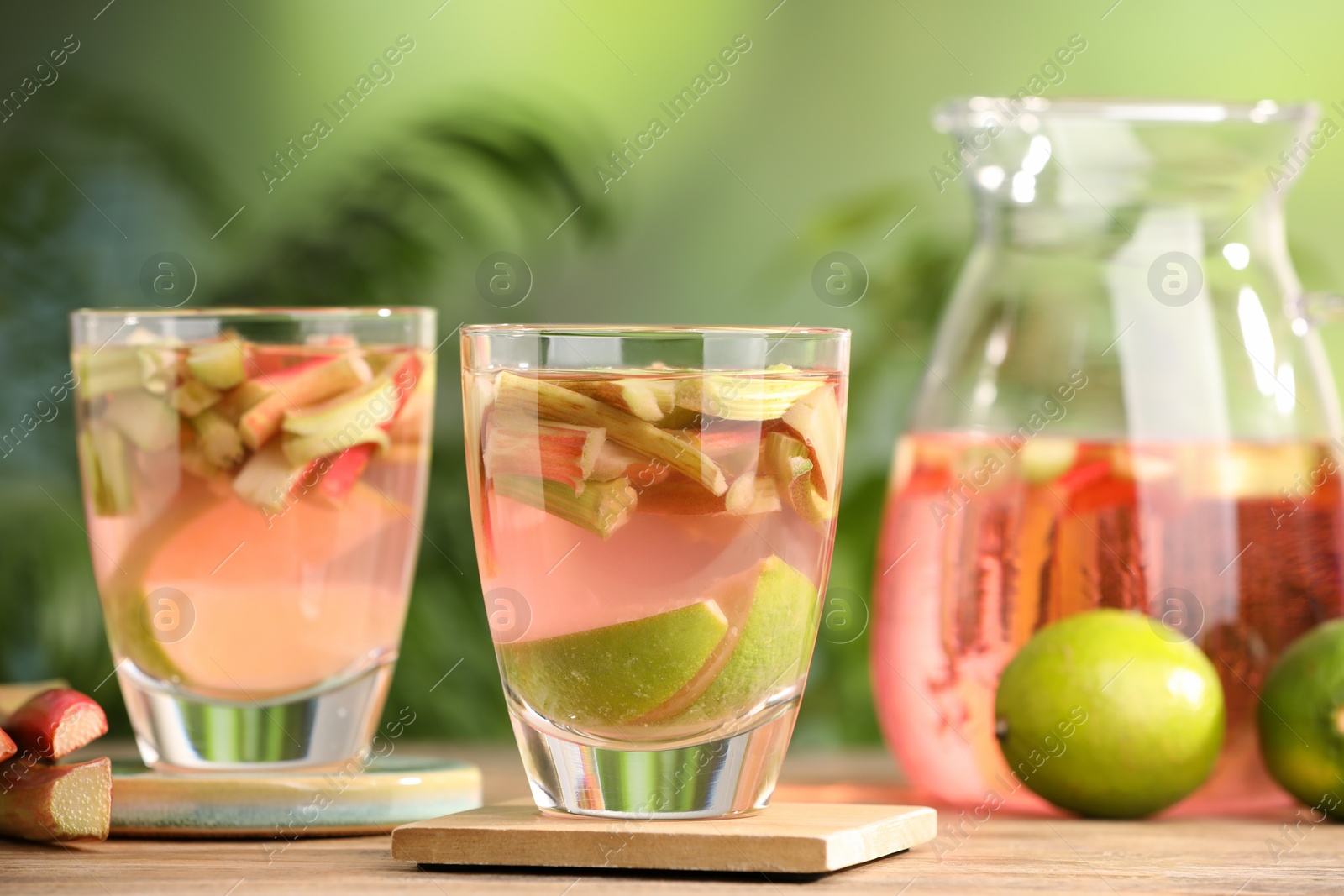 Photo of Glasses and jug of tasty rhubarb cocktail with lime fruits on wooden table outdoors