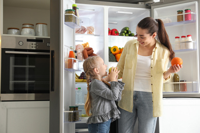 Young mother and her daughter with fruits near open refrigerator in kitchen