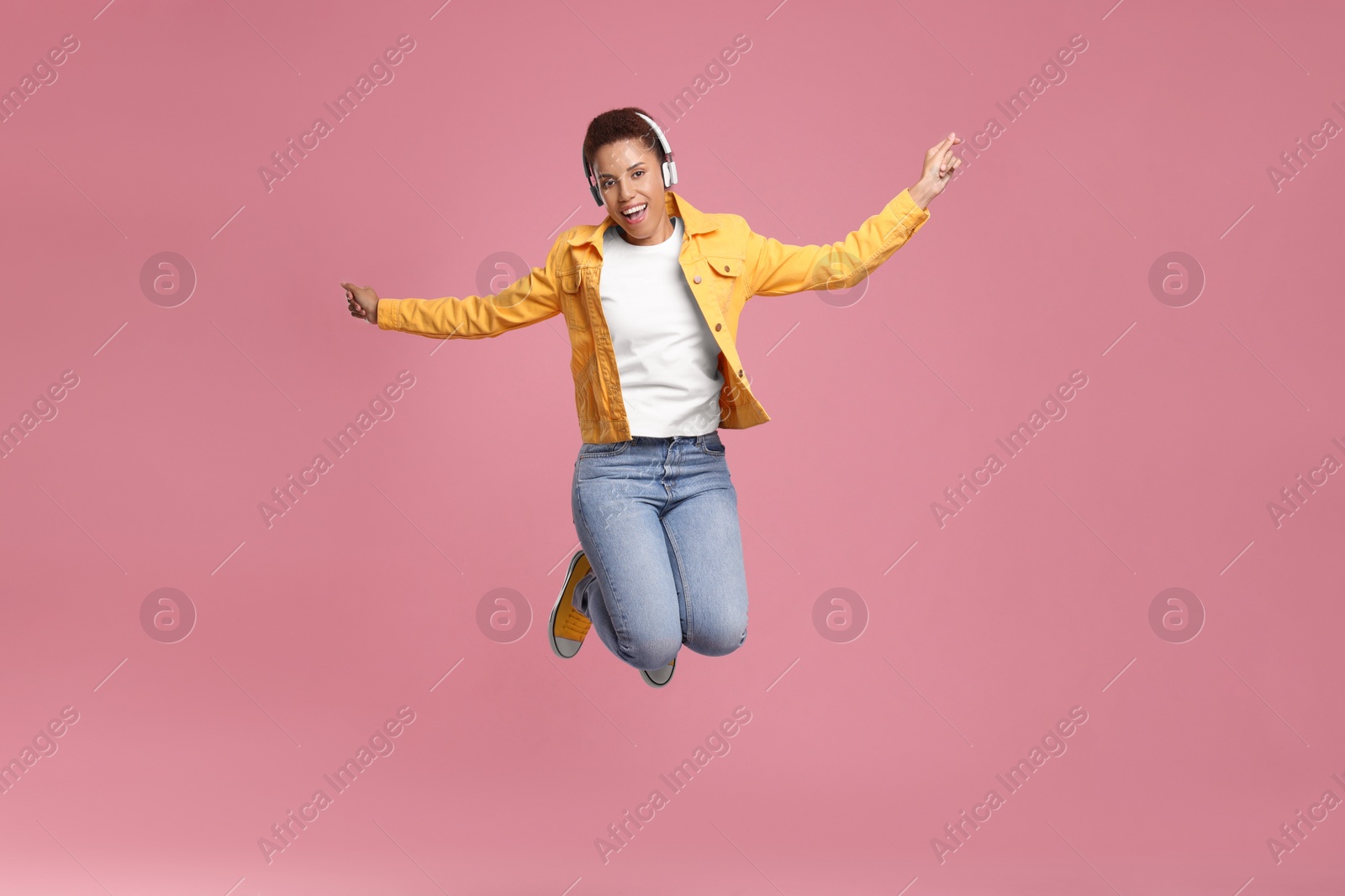 Photo of Happy young woman in headphones jumping while dancing on pink background