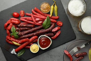 Photo of Different thin dry smoked sausages, sauces and glasses of beer on grey table, flat lay