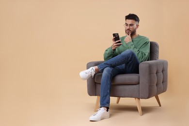 Photo of Handsome young man using smartphone in armchair on beige background, space for text