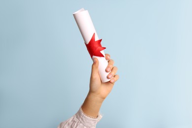 Photo of Student holding rolled diploma with red ribbon on light blue background, closeup
