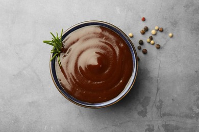 Photo of Tasty barbeque sauce in bowl, rosemary and peppercorns on grey textured table, top view