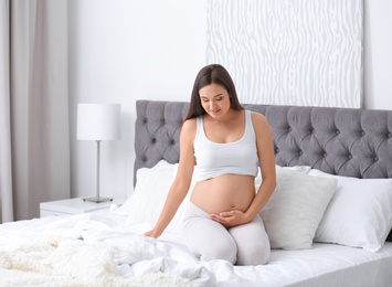 Photo of Young beautiful pregnant woman sitting on bed and touching her belly at home