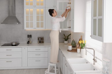 Photo of Woman on ladder putting white jar into cupboard at home