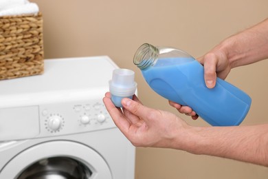 Photo of Man pouring fabric softener from bottle into cap near washing machine indoors, closeup