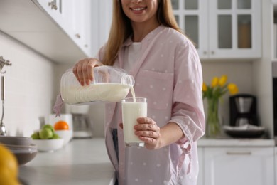 Young woman pouring milk from gallon bottle into glass near light countertop in kitchen, closeup