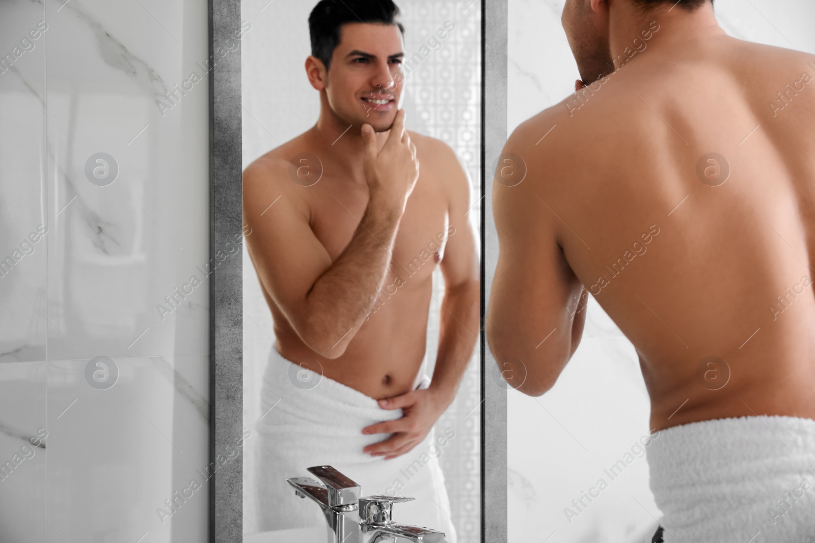 Photo of Handsome man touching his smooth face after shaving near mirror in bathroom