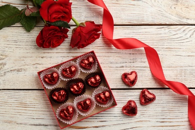 Photo of Heart shaped chocolate candies in box, roses and red ribbon on white wooden table, flat lay