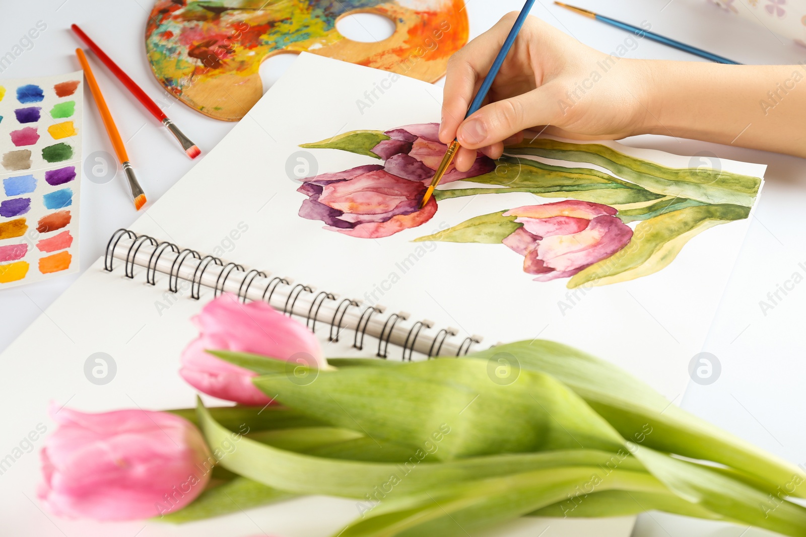 Photo of Woman painting tulips in sketchbook at white table, closeup