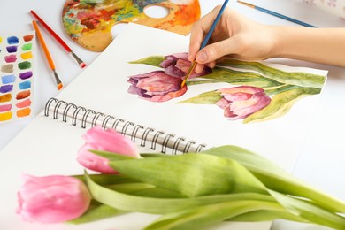 Photo of Woman painting tulips in sketchbook at white table, closeup