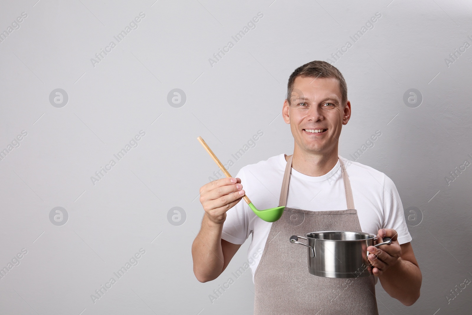 Photo of Happy man with cooking pot and ladle on light grey background. Space for text