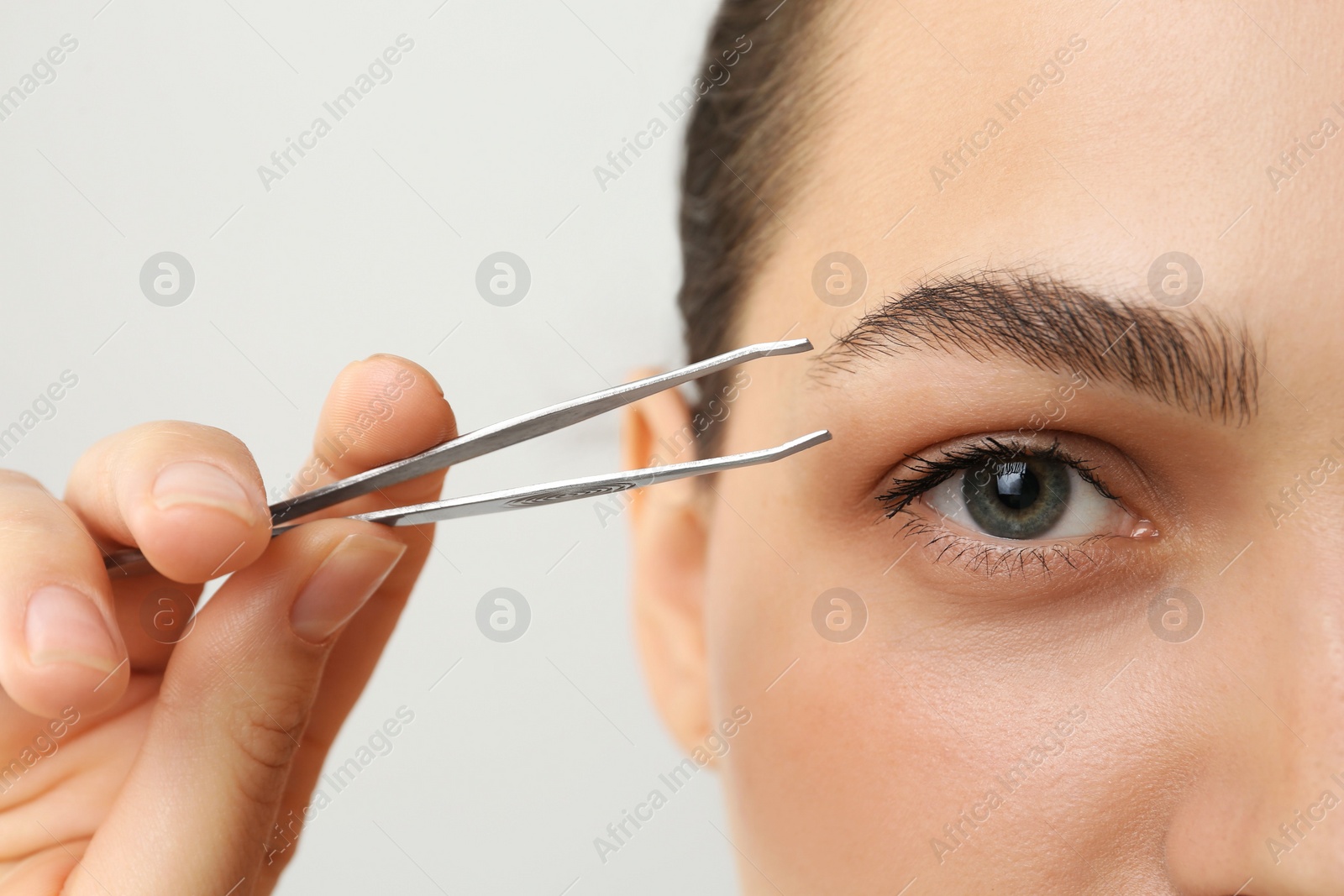 Photo of Eyebrow correction. Young woman with tweezers on light grey background, closeup