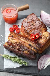 Photo of Pieces of baked pork belly served with sauce, rosemary and chili pepper on grey table