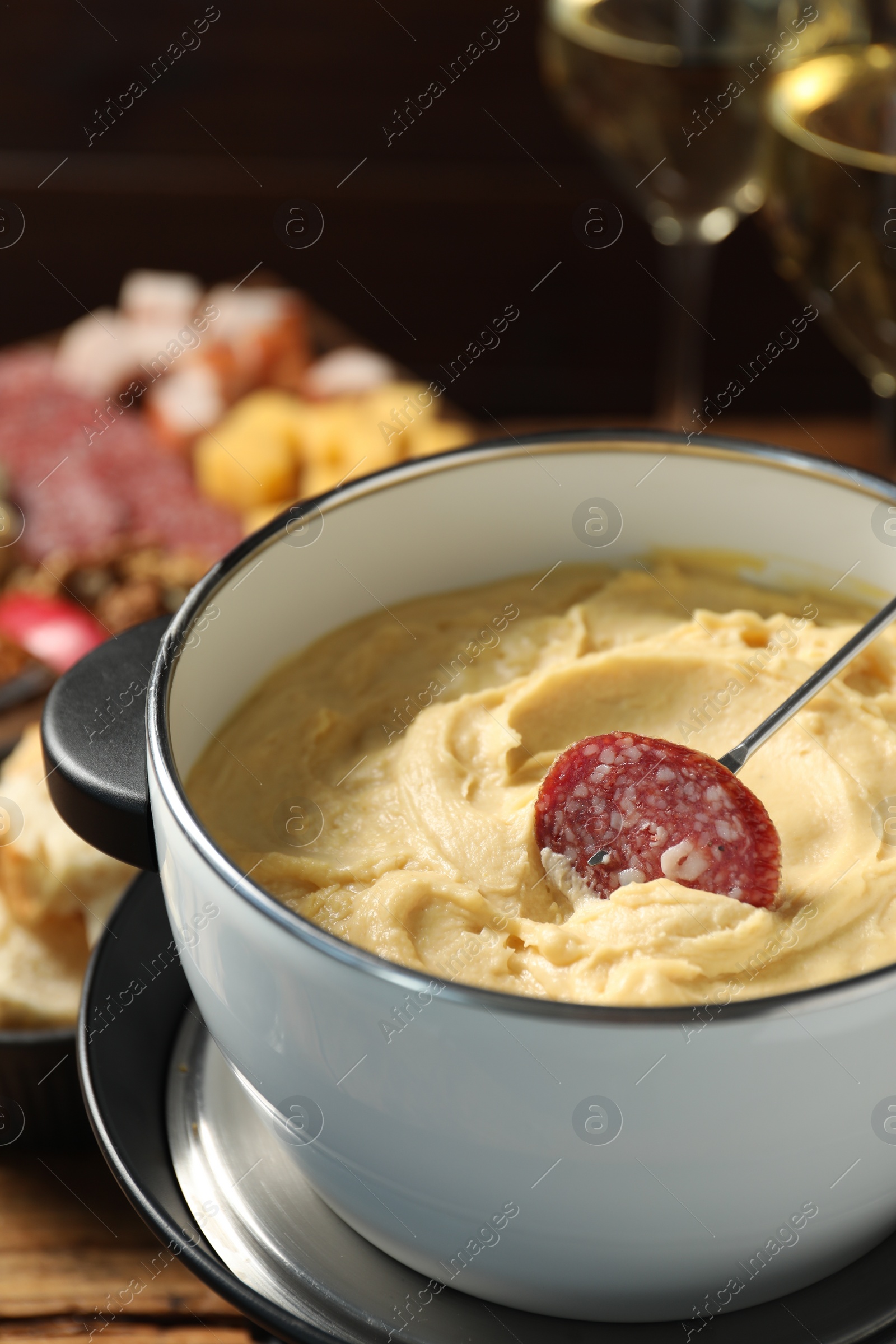 Photo of Dipping piece of sausage into fondue pot with melted cheese at table, closeup