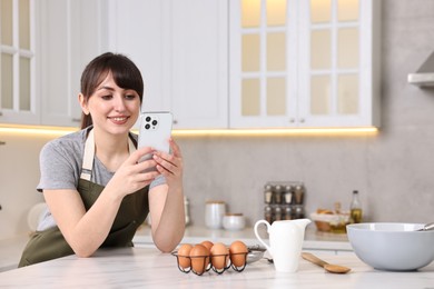 Photo of Happy young housewife using smartphone while cooking at white marble table in kitchen