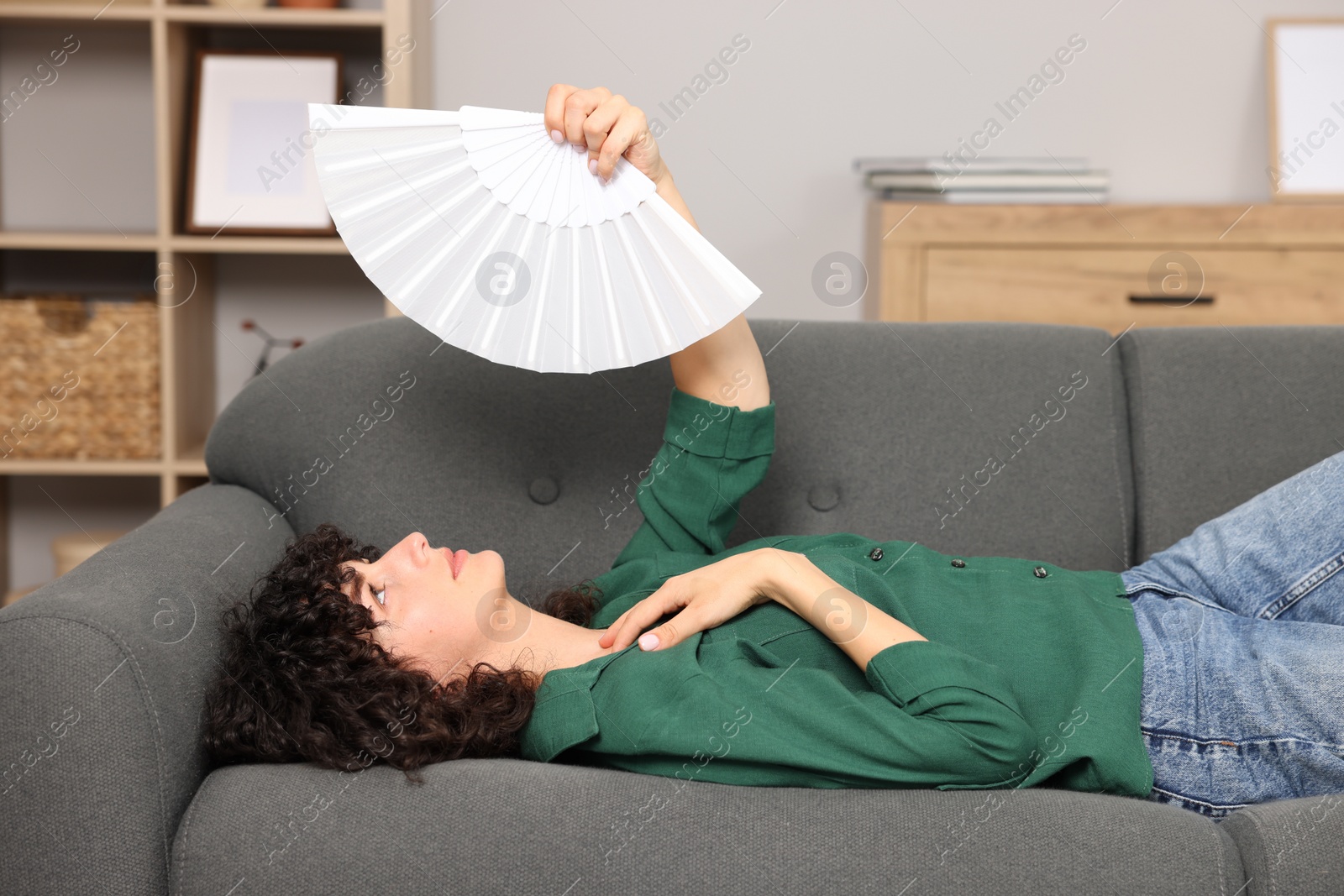 Photo of Young woman waving hand fan to cool herself on sofa at home