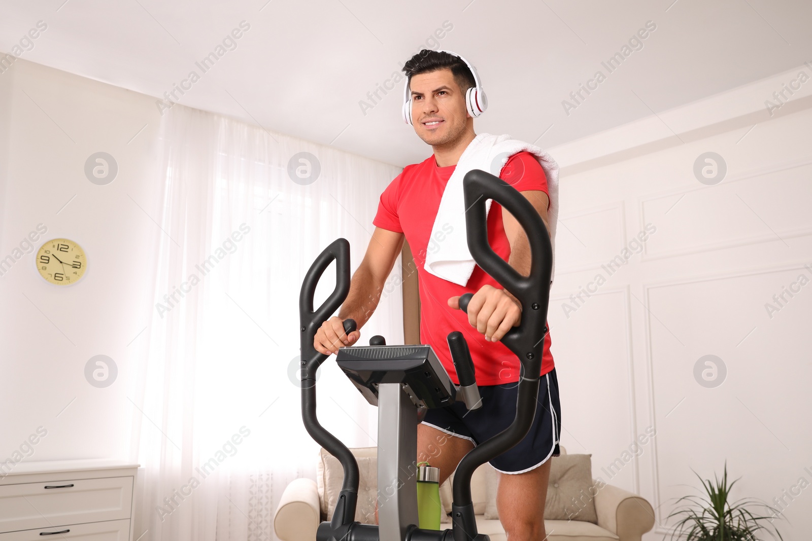 Photo of Man with headphones and towel using modern elliptical machine at home
