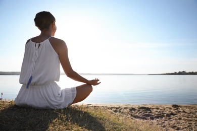 Young woman meditating near river at sunset, space for text. Nature healing power