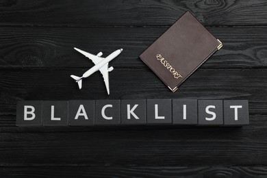Photo of Cubes with word Blacklist, toy airplane and passport on black wooden table, flat lay