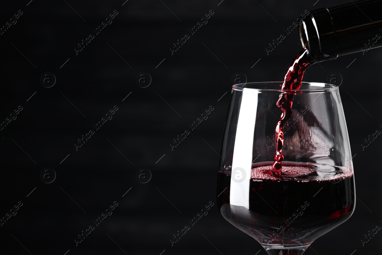 Photo of Pouring red wine into glass against dark background, closeup. Space for text