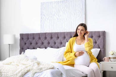 Photo of Young beautiful pregnant woman sitting on bed and touching her belly at home