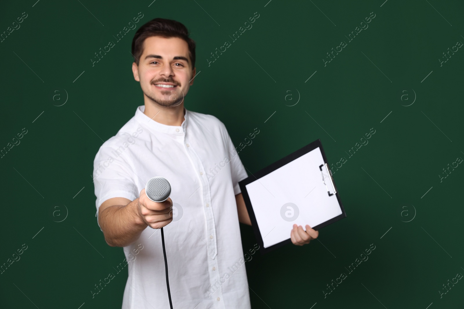 Photo of Young man in casual clothes holding microphone and clipboard on color background. Space for text