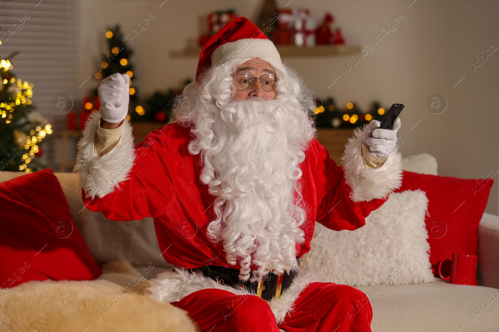 Photo of Merry Christmas. Santa Claus with remote control watching TV on sofa at home