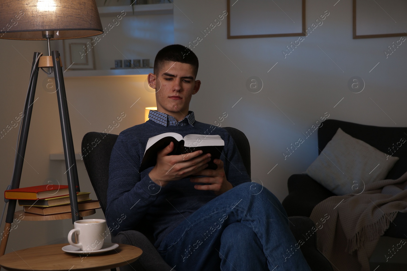 Photo of Young man with cup of drink reading book in cozy room at night