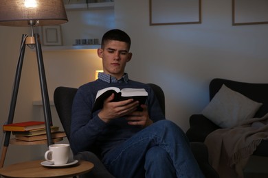Young man with cup of drink reading book in cozy room at night