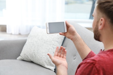 Young man using video chat on smartphone in living room, closeup. Space for design