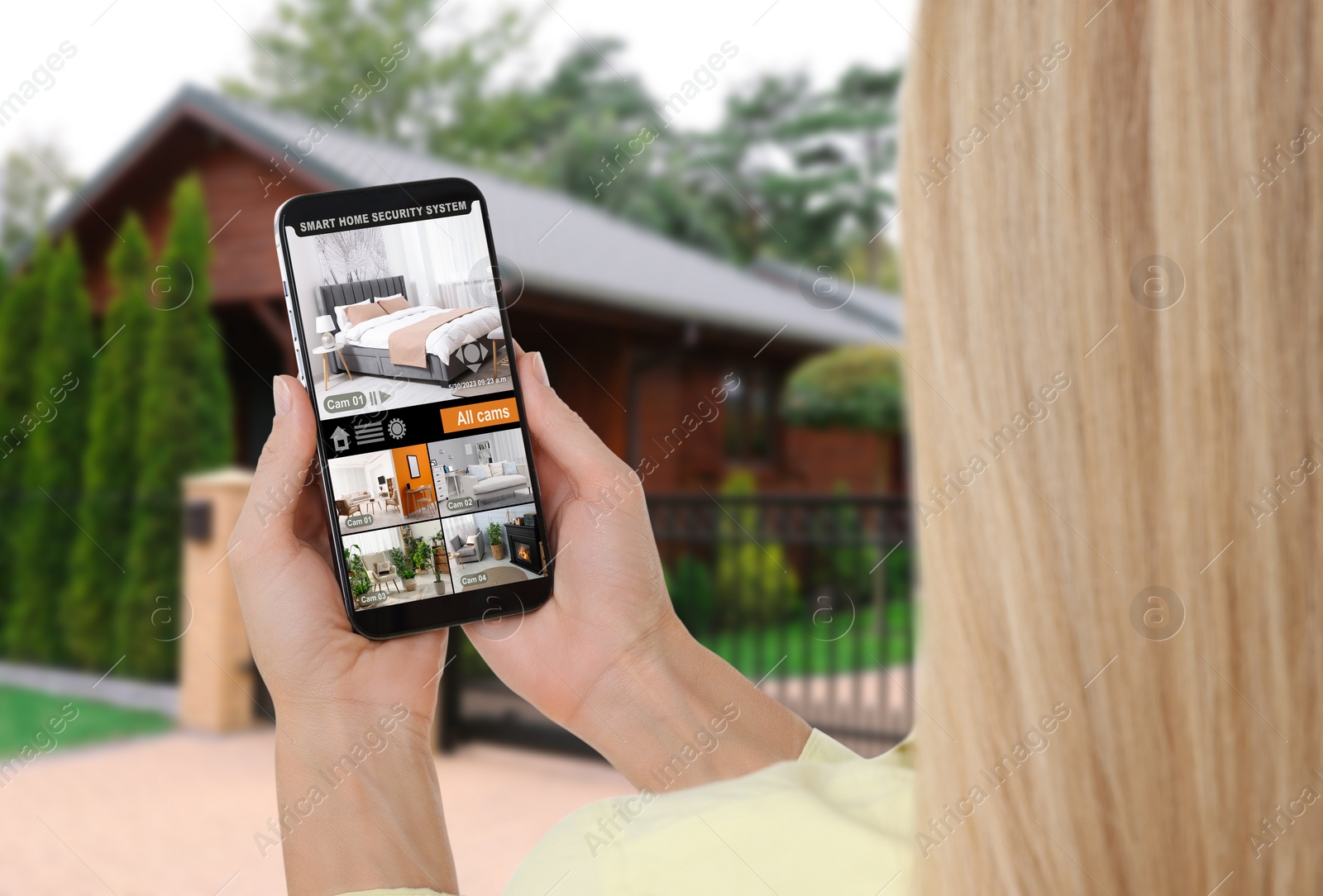 Image of Woman using smart home security system on mobile phone near house outdoors. Device showing different rooms through cameras