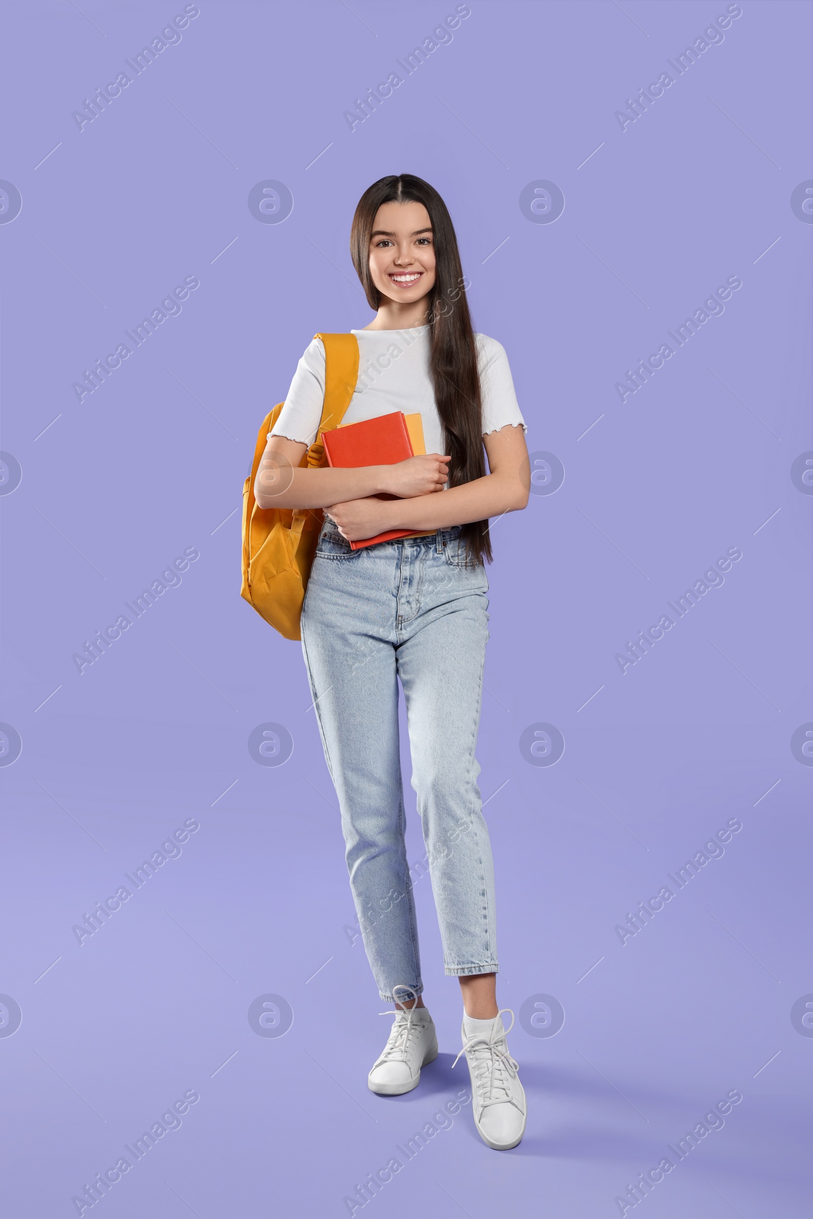 Photo of Happy teenage girl with backpack and textbooks on violet background