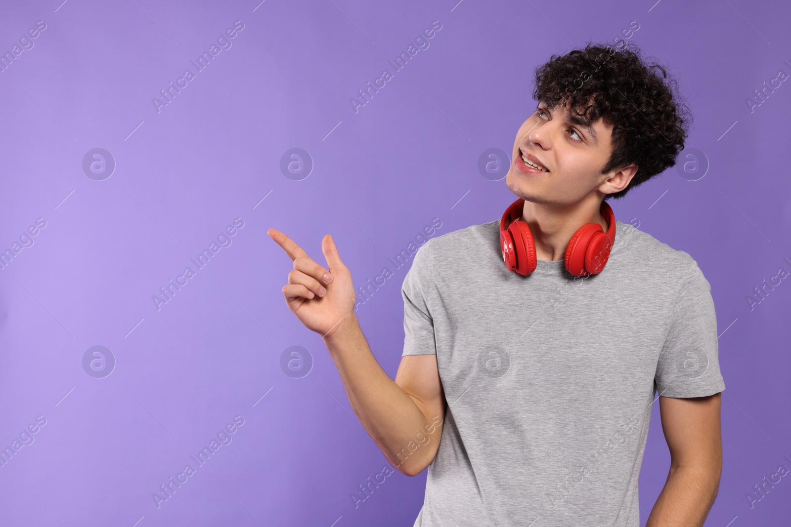 Photo of Handsome young man with headphones pointing at something on violet background. Space for text