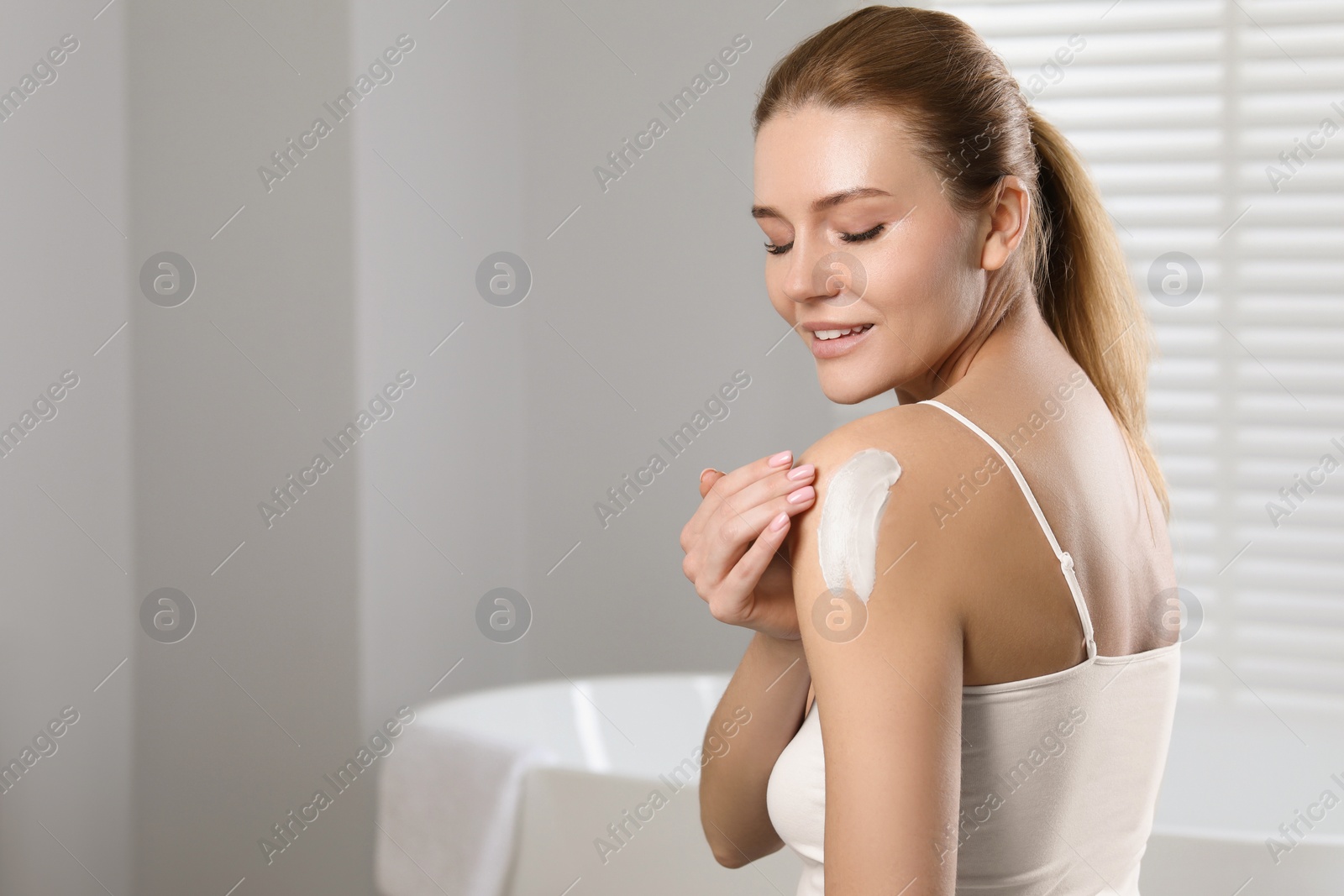 Photo of Happy woman applying body cream onto shoulder in bathroom, space for text
