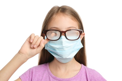 Photo of Little girl wiping foggy glasses caused by wearing disposable mask on white background. Protective measure during coronavirus pandemic