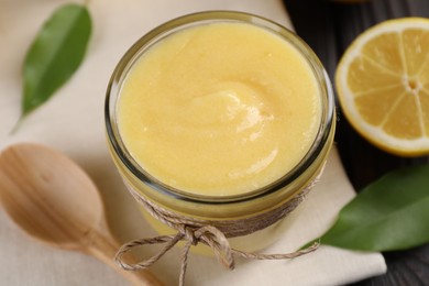 Photo of Delicious lemon curd in bowl, fresh citrus fruit, spoon and green leaves on table, closeup