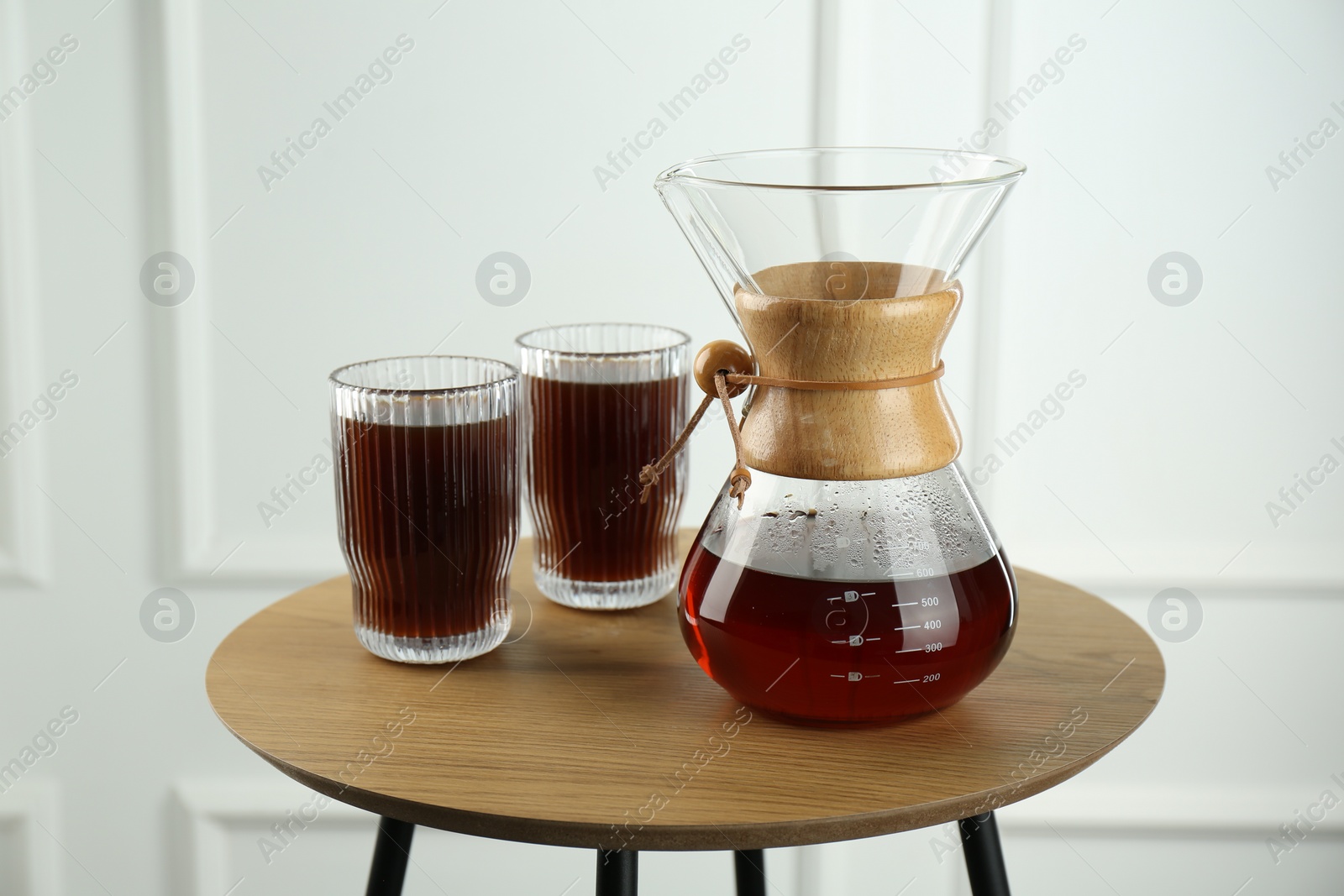 Photo of Glass chemex coffeemaker and glasses of coffee on wooden table against white wall