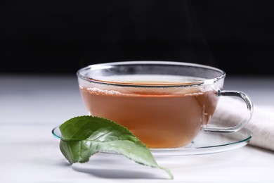 Photo of Aromatic hot tea in glass cup and leaf on white table against black background, closeup