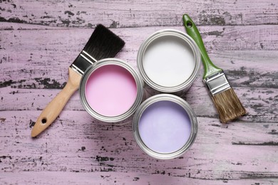 Photo of Cans of white, lilac and pink paints with brushes on color wooden table, flat lay