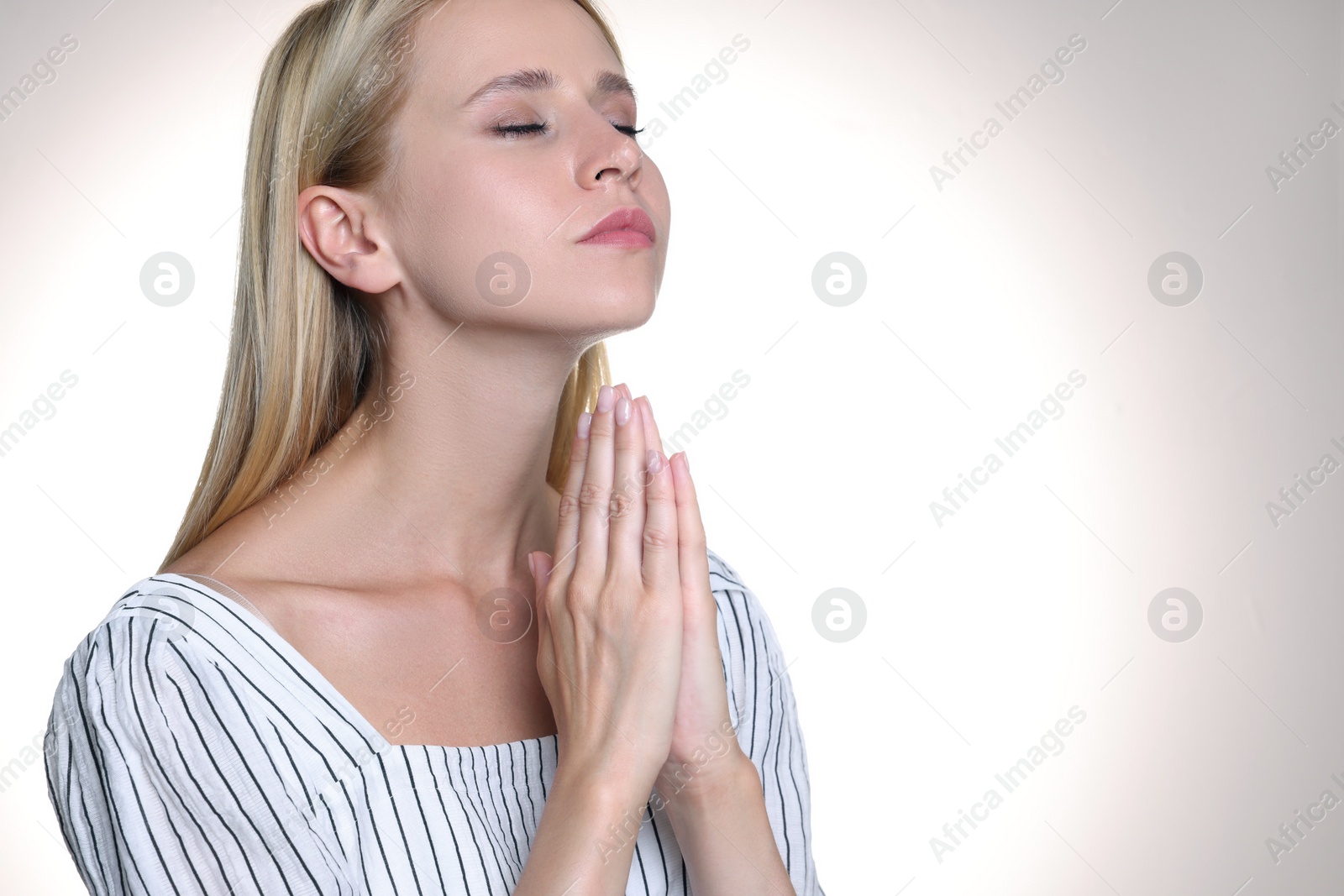 Photo of Religious young woman with clasped hands praying against light background. Space for text