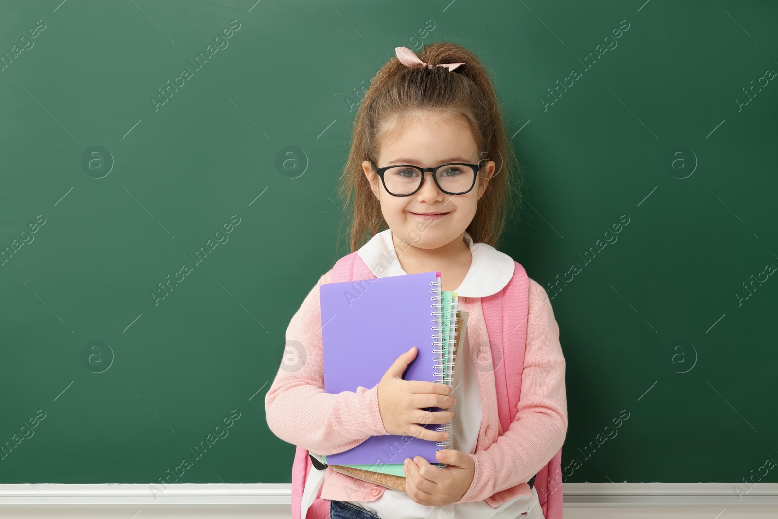 Photo of Happy little school child with notebooks near chalkboard