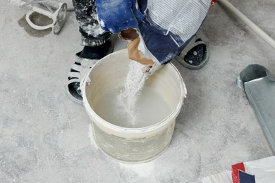 Photo of Professional worker pouring powdered plaster into bucket indoors, closeup