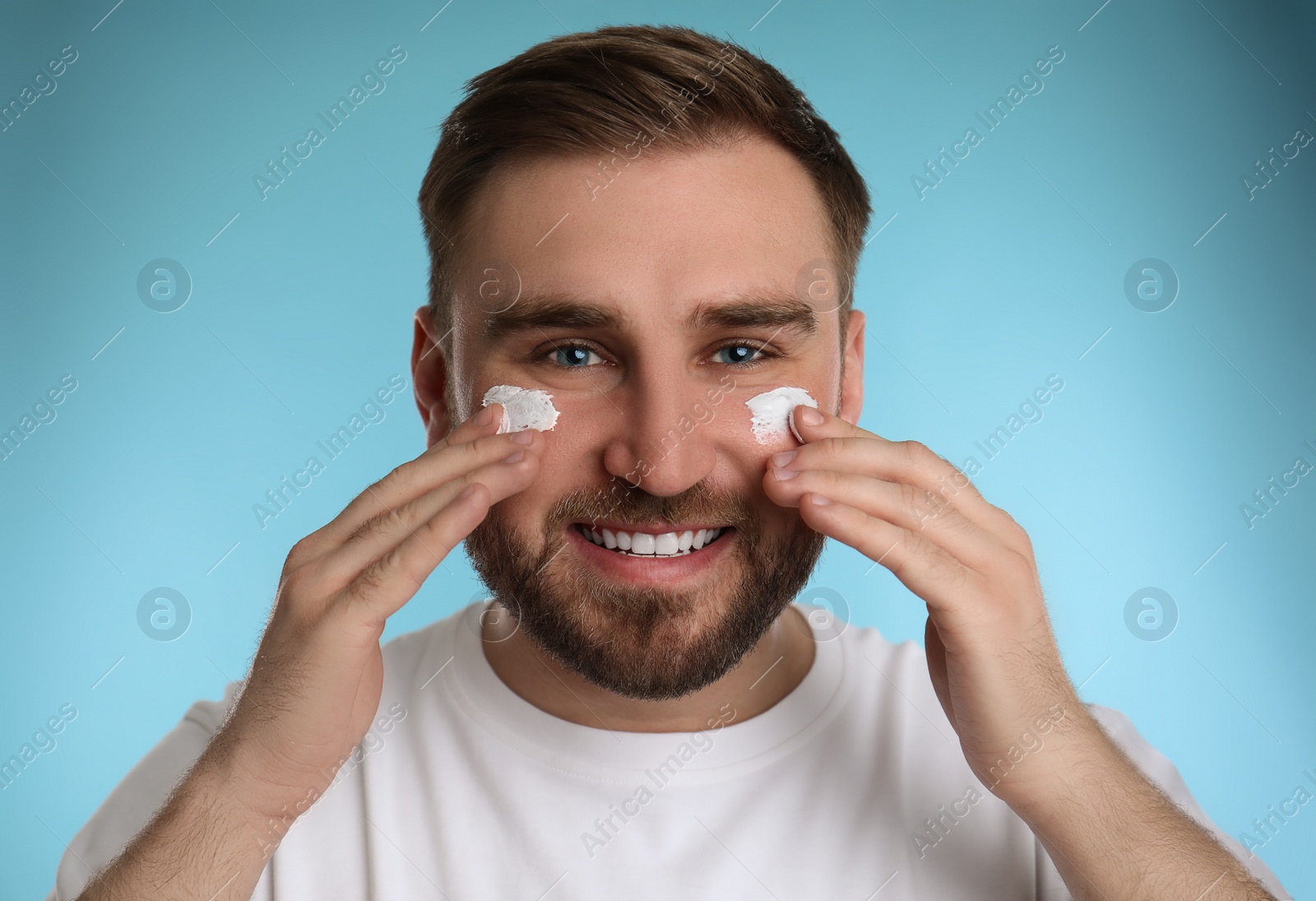 Photo of Happy young man applying facial cream on light blue background