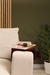 Photo of Magazine and remote control on sofa armrest wooden table in room, space for text. Interior element